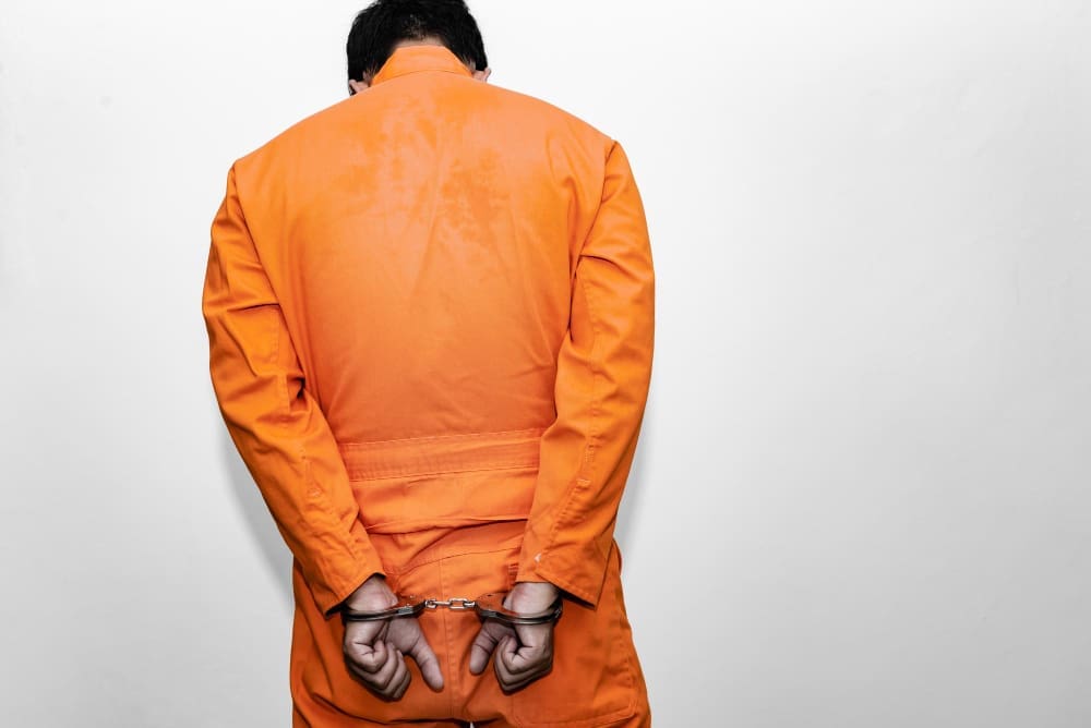 A person wearing a bright orange jumpsuit is seen from behind, with their hands cuffed behind their back, standing against a plain light-colored background.