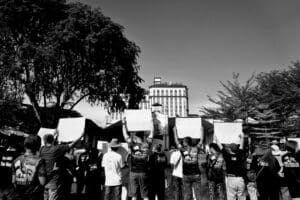 A black and white photograph of a crowd of protesters seen from behind. Many people are holding up blank white signs while facing a large building in the distance, framed by trees.