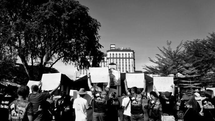 A black and white photograph of a crowd of protesters seen from behind. Many people are holding up blank white signs while facing a large building in the distance, framed by trees.