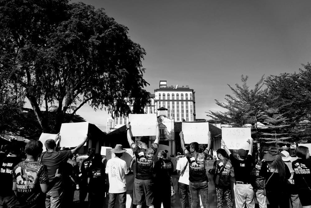 A black and white photograph of a crowd of protesters seen from behind. Many people are holding up blank white signs while facing a large building in the distance, framed by trees.