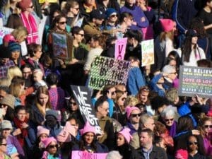 A dense crowd of diverse people at a protest, many wearing pink knitted "pussy hats." They are holding up handmade signs with political slogans, including "Grab 'em By The Midterms" and another listing progressive beliefs like "Black Lives Matter" and "Women's Rights Are Human Rights."