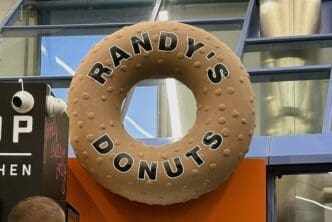A giant, iconic brown donut-shaped sign for "Randy's Donuts" with raised "sprinkles" and the brand name in black letters, hanging in a modern indoor space at an airport.