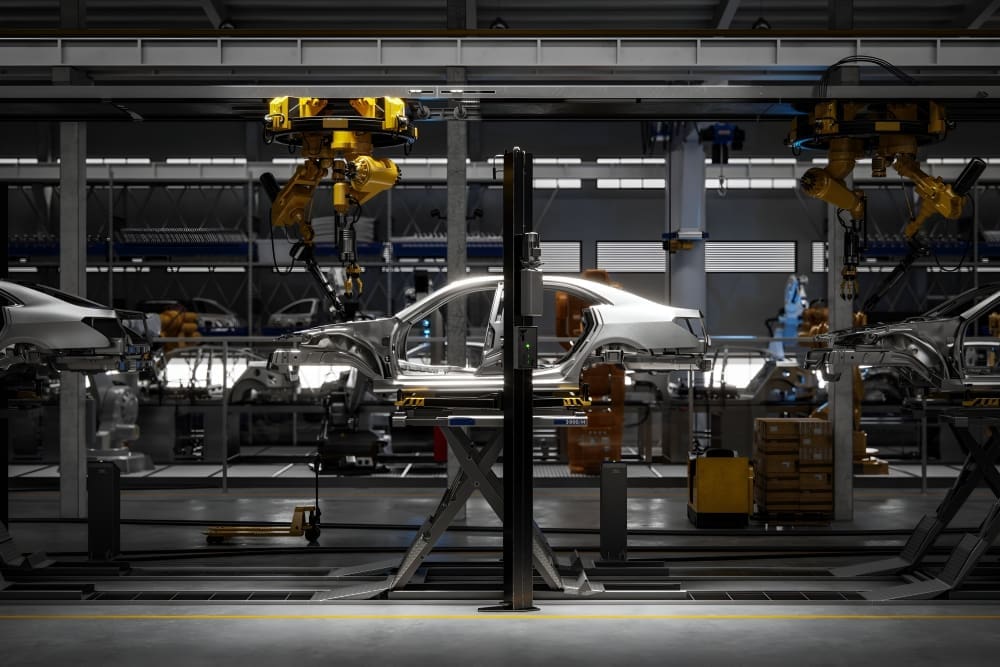The metal frame of a car is held on a lift in the center of a factory assembly line, surrounded by large, yellow robotic arms used for welding and manufacturing.