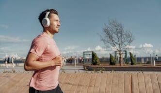 A man who appears to be White is jogging outdoors in an urban setting, wearing a pink t-shirt, dark shorts, and large white over-ear headphones. He looks focused, with a blurred background of people and greenery.