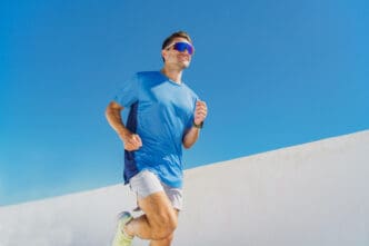 A man in a blue running shirt and light shorts is seen in mid-stride, wearing large, mirrored performance running sunglasses, running outdoors against a clear blue sky and a white wall.