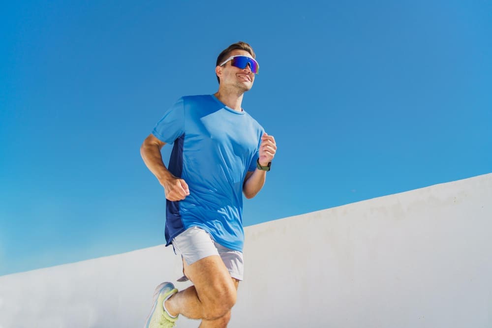 A man in a blue running shirt and light shorts is seen in mid-stride, wearing large, mirrored performance running sunglasses, running outdoors against a clear blue sky and a white wall.