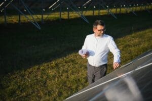 A middle-aged male engineer or business owner, who appears to be White and is wearing glasses, stands in a field of solar panels, holding blueprints while inspecting a panel's surface during golden hour.