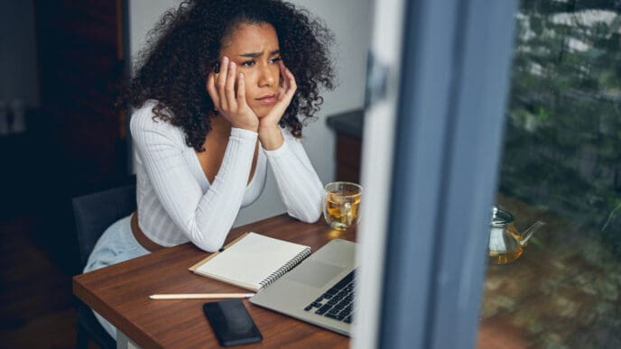 A young woman, who appears to be Black, sits with a sad expression in casual clothes. She is holding her head in her hands, conveying a sense of distress or sadness.