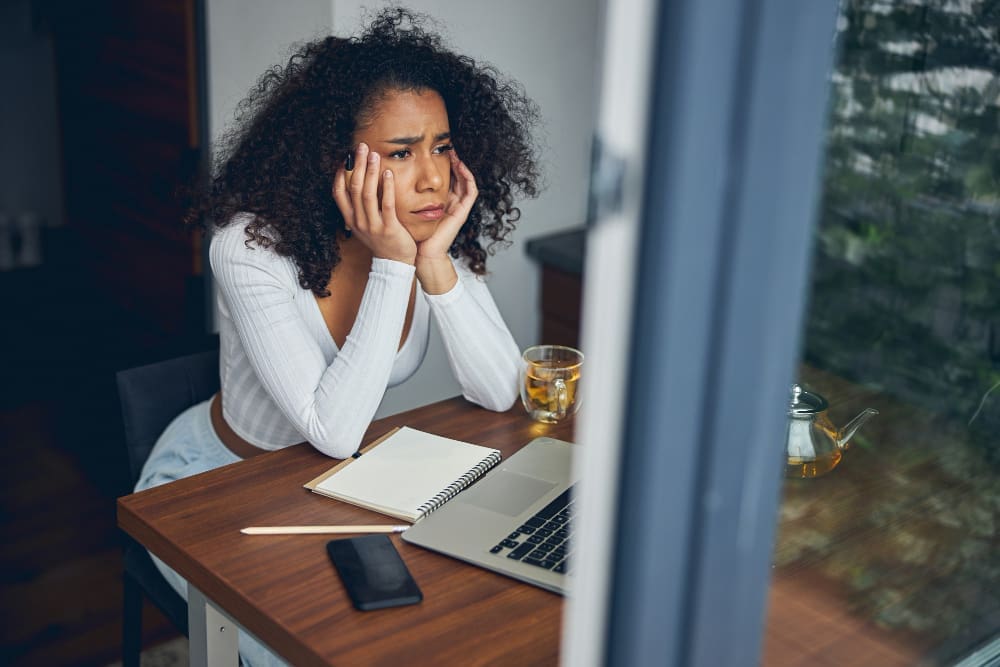 A young woman, who appears to be Black, sits with a sad expression in casual clothes. She is holding her head in her hands, conveying a sense of distress or sadness.