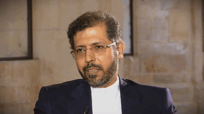 A professional portrait of Saeed Khatibzadeh, an Iranian diplomat with dark hair, a beard, and glasses, wearing a dark suit and white shirt, appearing to be speaking during an interview.