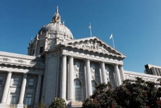 A low-angle view of San Francisco City Hall, showcasing its grand white dome with a finial, large classical columns, and ornate sculptures on its pediment, symbolizing civic governance and justice under a clear blue sky.