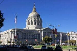 A wide-angle, daytime view of San Francisco City Hall, showcasing its grand white dome with a golden finial, classical columns, and an American flag flying prominently in front of it under a clear blue sky.