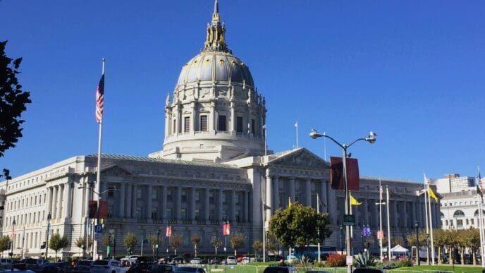 A wide-angle, daytime view of San Francisco City Hall, showcasing its grand white dome with a golden finial, classical columns, and an American flag flying prominently in front of it under a clear blue sky.