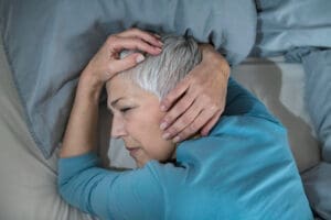 A woman who appears to be White, with short gray hair, lies on her side in bed with her hands on her head, looking distressed or in pain, symbolizing the struggle with sleep deprivation.