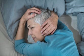 A woman who appears to be White, with short gray hair, lies on her side in bed with her hands on her head, looking distressed or in pain, symbolizing the struggle with sleep deprivation.