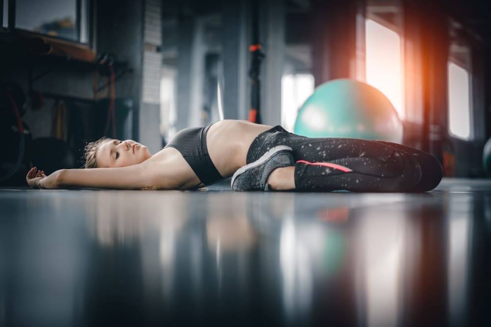 A young woman in athletic wear lies flat on her back on a gym floor with her arms stretched out to the sides and legs bent, eyes closed, in a pose suggesting deep relaxation or meditation.