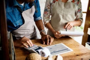 A close-up view of two small business owners, a man and a woman, working behind a wooden counter. The man is using a calculator, and the woman is holding a clipboard, with a digital tablet showing an order form between them.