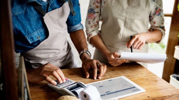 A close-up view of two small business owners, a man and a woman, working behind a wooden counter. The man is using a calculator, and the woman is holding a clipboard, with a digital tablet showing an order form between them.