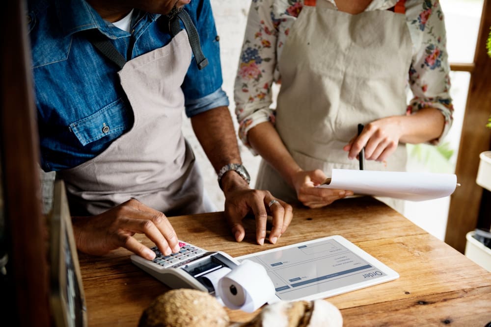 A close-up view of two small business owners, a man and a woman, working behind a wooden counter. The man is using a calculator, and the woman is holding a clipboard, with a digital tablet showing an order form between them.