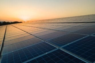 An elevated view looking across a vast field of solar panels at a solar farm. The warm, golden light of the setting or rising sun is visible on the horizon and reflects off the panel surfaces.