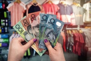 A close-up of a person's hands holding a fan of Australian dollar banknotes ($100, $20, and $10 notes) inside a clothing store, with racks of clothes blurred in the background.