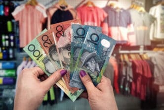 A close-up of a person's hands holding a fan of Australian dollar banknotes ($100, $20, and $10 notes) inside a clothing store, with racks of clothes blurred in the background.