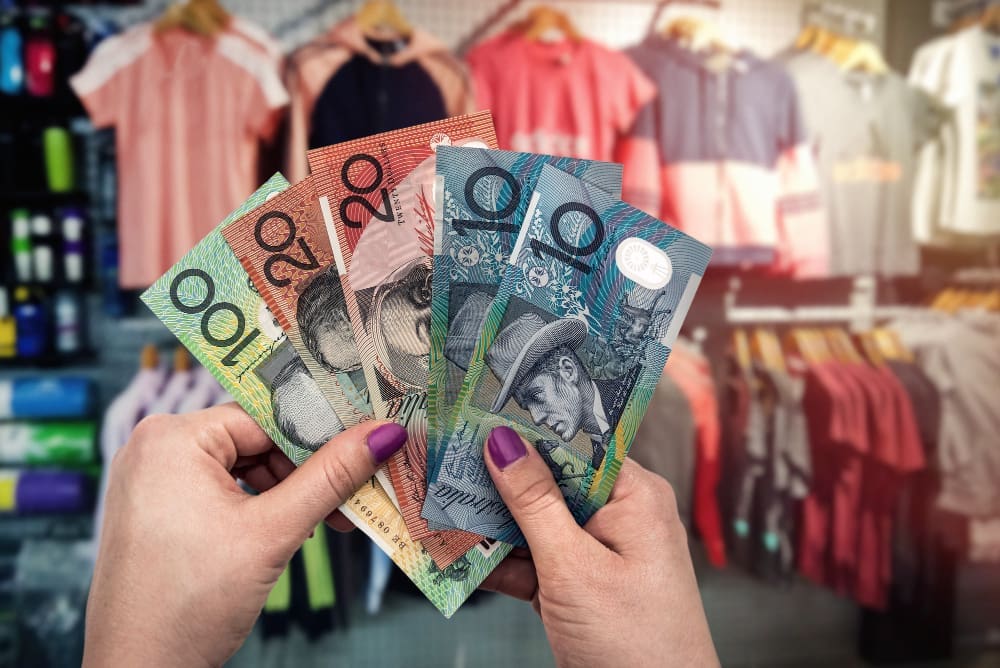 A close-up of a person's hands holding a fan of Australian dollar banknotes ($100, $20, and $10 notes) inside a clothing store, with racks of clothes blurred in the background.