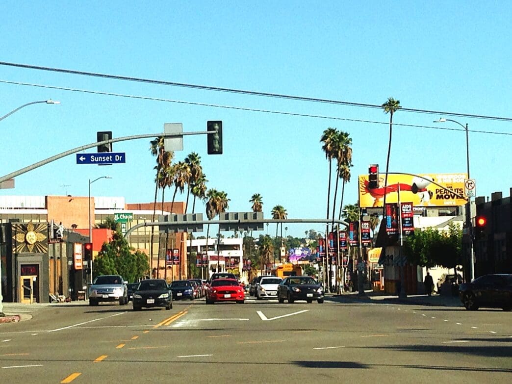 A daytime, street-level view of a wide avenue in Los Angeles, California, lined with palm trees. A street sign in the image clearly indicates the location is Sunset Drive.