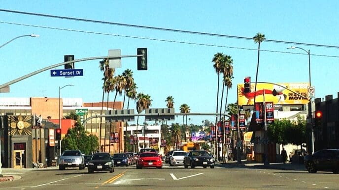 A daytime, street-level view of a wide avenue in Los Angeles, California, lined with palm trees. A street sign in the image clearly indicates the location is Sunset Drive.