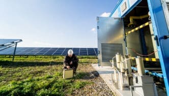 An engineer wearing a hard hat crouches to work on testing equipment at a solar farm, with rows of solar panels in the background and an open energy storage container unit nearby.