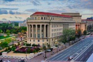 An elevated photograph of the historic, neoclassical Apex Building, the headquarters of the Federal Trade Commission in Washington, D.C., viewed at dusk from across a wide avenue.