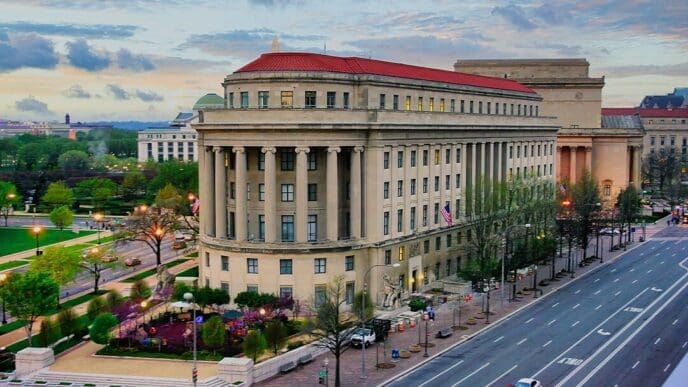 An elevated photograph of the historic, neoclassical Apex Building, the headquarters of the Federal Trade Commission in Washington, D.C., viewed at dusk from across a wide avenue.