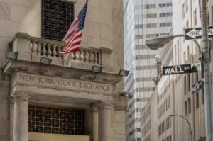 A street-level view of the New York Stock Exchange building on Wall Street in New York City. The image shows the building's neoclassical facade, an American flag, and the "WALL ST" street sign.