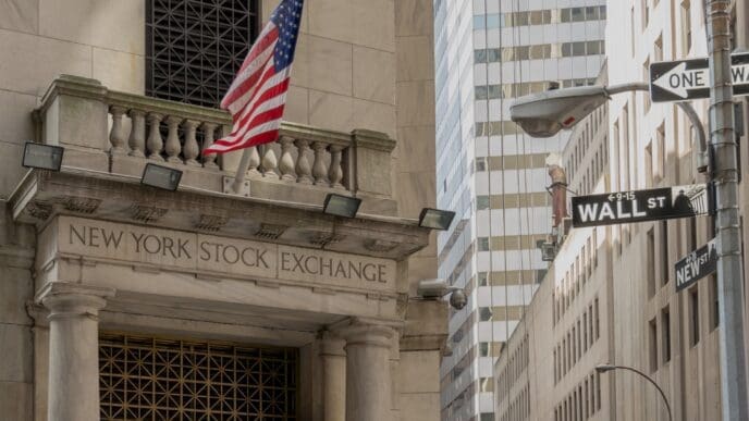 A street-level view of the New York Stock Exchange building on Wall Street in New York City. The image shows the building's neoclassical facade, an American flag, and the "WALL ST" street sign.