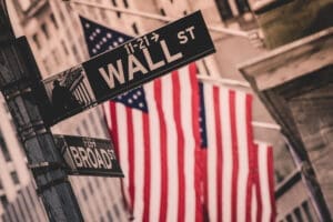 A close-up, stylized photograph of the "WALL ST" street sign in New York City, with the "BROAD ST" sign below it. In the blurred background, American flags hang on the facade of the New York Stock Exchange.