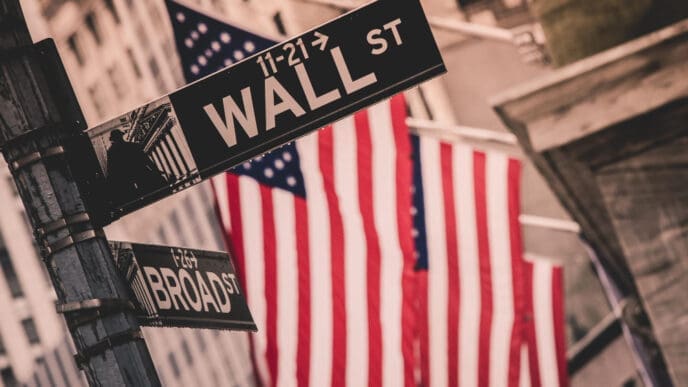 A close-up, stylized photograph of the "WALL ST" street sign in New York City, with the "BROAD ST" sign below it. In the blurred background, American flags hang on the facade of the New York Stock Exchange.