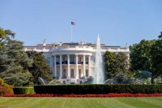 The White House's iconic South Portico is seen on a sunny day, with its white neoclassical facade, central columns, and the U.S. flag flying above. A fountain sprays water on the green lawn in the foreground, bordered by red flowers.