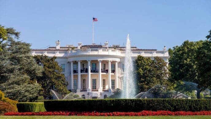 The White House's iconic South Portico is seen on a sunny day, with its white neoclassical facade, central columns, and the U.S. flag flying above. A fountain sprays water on the green lawn in the foreground, bordered by red flowers.