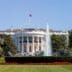 The White House's iconic South Portico is seen on a sunny day, with its white neoclassical facade, central columns, and the U.S. flag flying above. A fountain sprays water on the green lawn in the foreground, bordered by red flowers.
