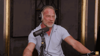 Todd Vande Hei, a man with graying hair and a beard, wears over-ear headphones and speaks into a professional podcast microphone in a studio setting.