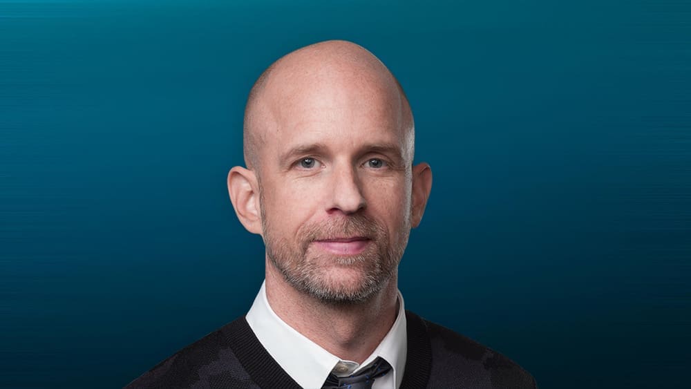 A professional headshot of Tor Myhren, a bald man with a short beard, wearing a sweater vest over a collared shirt and tie. He is looking directly at the camera against a textured blue background.