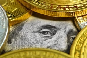 A close-up photograph of a U.S. hundred-dollar bill, showing Benjamin Franklin's eye, surrounded and partially covered by several gold and silver cryptocurrency coins.