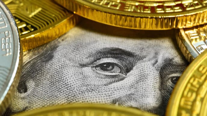 A close-up photograph of a U.S. hundred-dollar bill, showing Benjamin Franklin's eye, surrounded and partially covered by several gold and silver cryptocurrency coins.