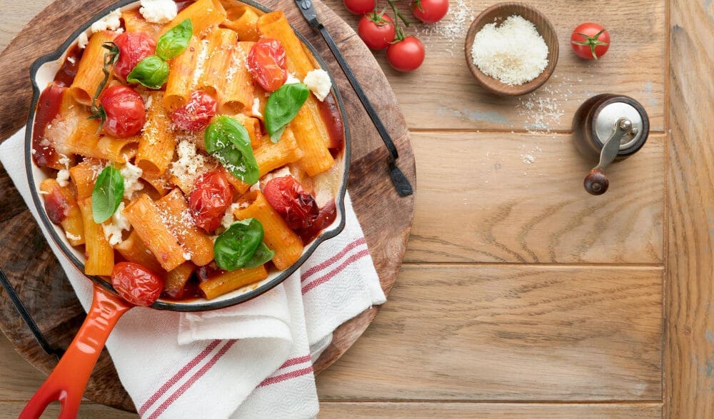 A top-down view of pasta tortiglioni with tomato sauce, baked cherry tomatoes, mozzarella cheese, and fresh basil in an orange skillet on a rustic wooden table, with grated Parmesan and whole tomatoes on the side.
