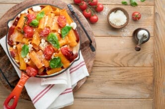 A top-down view of pasta tortiglioni with tomato sauce, baked cherry tomatoes, mozzarella cheese, and fresh basil in an orange skillet on a rustic wooden table, with grated Parmesan and whole tomatoes on the side.