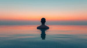 A person is seen from behind, submerged chest-deep in a calm body of water (like an infinity pool or ocean) at sunrise or sunset, facing a serene horizon with a sky gradient of blue, orange, and pink, symbolizing meditation.