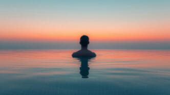 A person is seen from behind, submerged chest-deep in a calm body of water (like an infinity pool or ocean) at sunrise or sunset, facing a serene horizon with a sky gradient of blue, orange, and pink, symbolizing meditation.