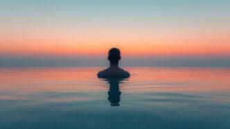 A person is seen from behind, submerged chest-deep in a calm body of water (like an infinity pool or ocean) at sunrise or sunset, facing a serene horizon with a sky gradient of blue, orange, and pink, symbolizing meditation.