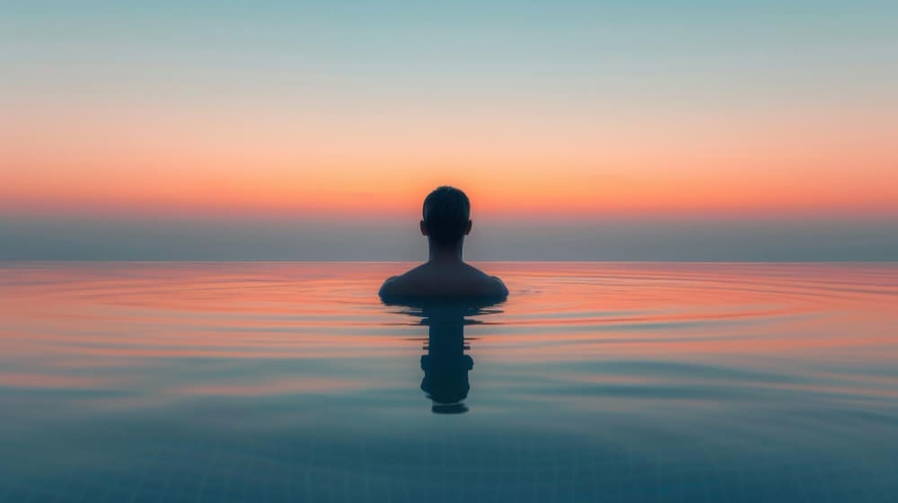 A person is seen from behind, submerged chest-deep in a calm body of water (like an infinity pool or ocean) at sunrise or sunset, facing a serene horizon with a sky gradient of blue, orange, and pink, symbolizing meditation.