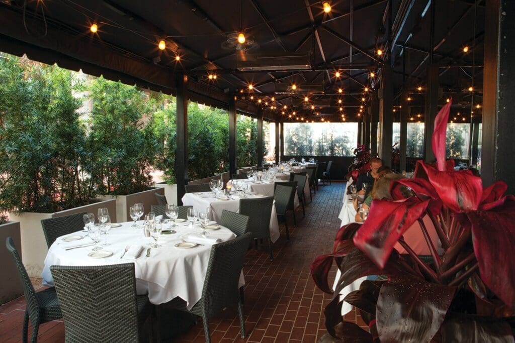 An elegantly set outdoor dining terrace of a restaurant, with numerous tables covered in white tablecloths under a dark canopy illuminated by string lights and surrounded by lush greenery.